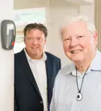 Persoonsalarm Thuis - oudere man met alarmknop om de nek, man op achtergrond, allebei naar alarmapparaat