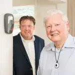 Persoonsalarm Thuis - oudere man met alarmknop om de nek, man op achtergrond, allebei naar alarmapparaat