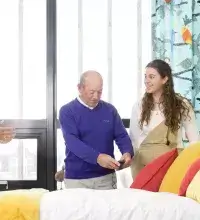 oudere man staat met dochter en gastvrouw bij een bed in Slimste Huis