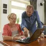 veilig online - senior vrouw zit met laptop aan tafel en haar man staat ernaast