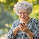 Oudere vrouw met bril en hoortoestel kijkt lachend op haar smartphone