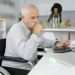 Oudere man in rolstoel met rekenmachine en laptop, hulp op de achtergrond