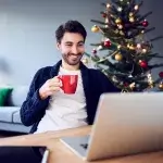 Een man zit achter een laptop met een kerstboom op de achtergrond.