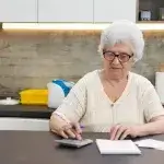 Een dame zit aan de keukentafel met rekenmachine, pen en papier en administratie